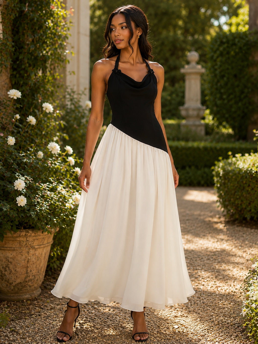 A woman wearing a two-tone maxi dress with a black cowl-neck bodice and a flowing white skirt, standing in a sunlit garden courtyard.