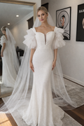 A woman wearing a white off-the-shoulder mermaid-style wedding gown with a sweetheart neckline, ruffled puff sleeves, and a long sheer veil, standing indoors with a mirror showing her reflection.