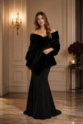 A woman stands in an elegant indoor room wearing a black off-the-shoulder faux-fur wrap draped over a floor-length black gown. The setting features warm wall paneling, a lit wall sconce, and a marble floor.