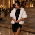 A woman stands in a grand indoor lobby wearing a voluminous white faux-fur jacket over a fitted black dress. The setting features marble stairs, warm candle lighting, and floral arrangements in the background.
