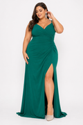 A woman modeling a floor-length emerald green gown with a V-neckline, ruched bodice, and high front slit, photographed against a plain light gray studio background.