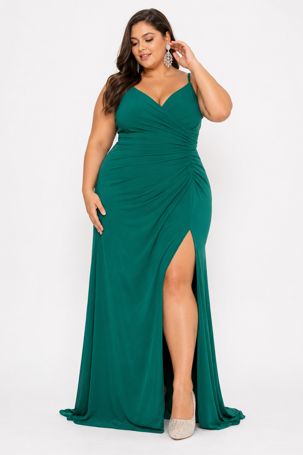 A woman modeling a floor-length emerald green gown with a V-neckline, ruched bodice, and high front slit, photographed against a plain light gray studio background.