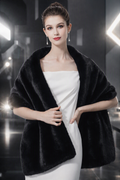 A woman modeling a black faux fur shawl draped over her shoulders, worn over a white strapless dress in a dark, modern studio setting with bright light flares.