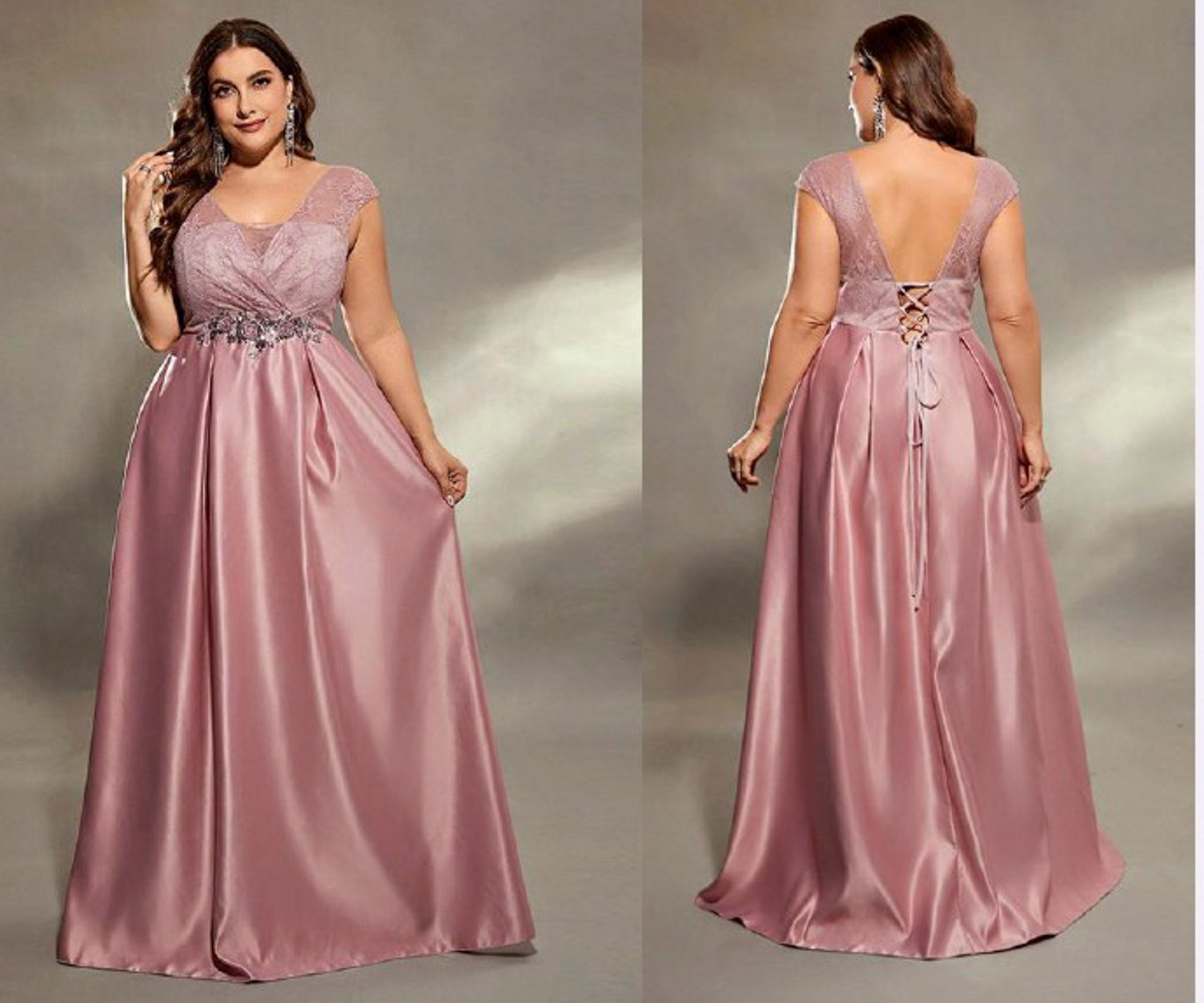 A plus-size model is shown wearing a dusty pink floor-length formal gown from the front and back against a neutral studio backdrop. The dress features a lace V-neck bodice, a beaded waistband, and an open lace-up back.