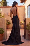 A woman wearing a floor-length black satin gown with a dramatic open back and draped train, photographed outdoors on a sunlit patio with stucco walls and potted plants.