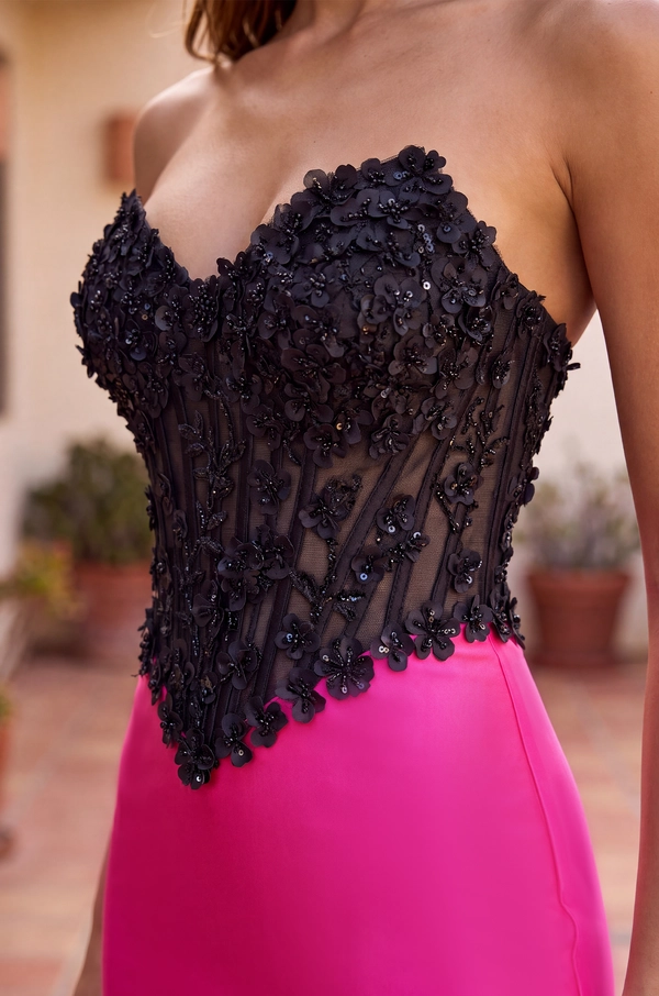 A close-up of a strapless black corset-style top with floral lace applique and sheer mesh panels, worn with a bright pink skirt in an outdoor setting.